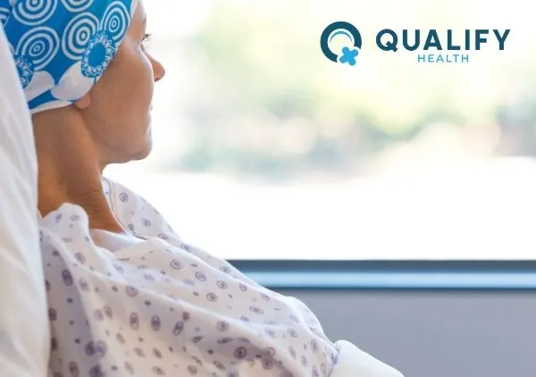 A medical patient gazing out a window, dressed in a dotted hospital gown and blue head covering.