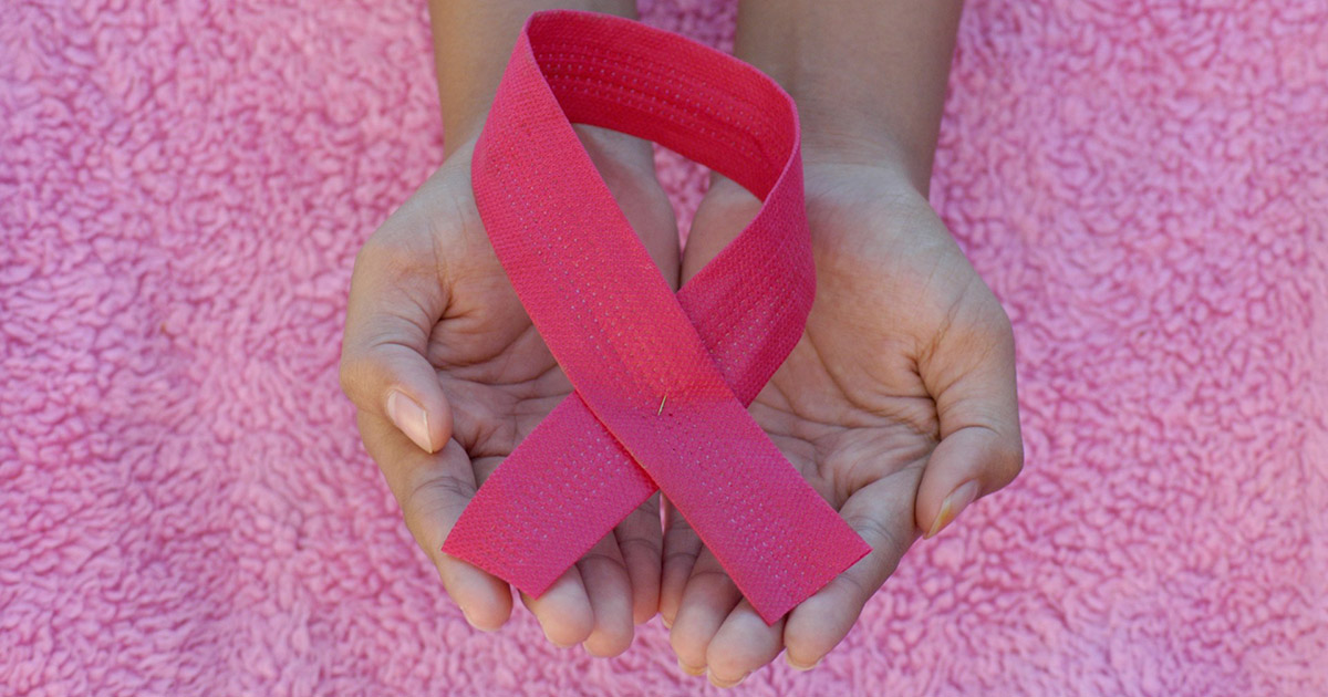 Hands gently holding a pink ribbon against a textured pink fabric background, symbolizing breast cancer awareness